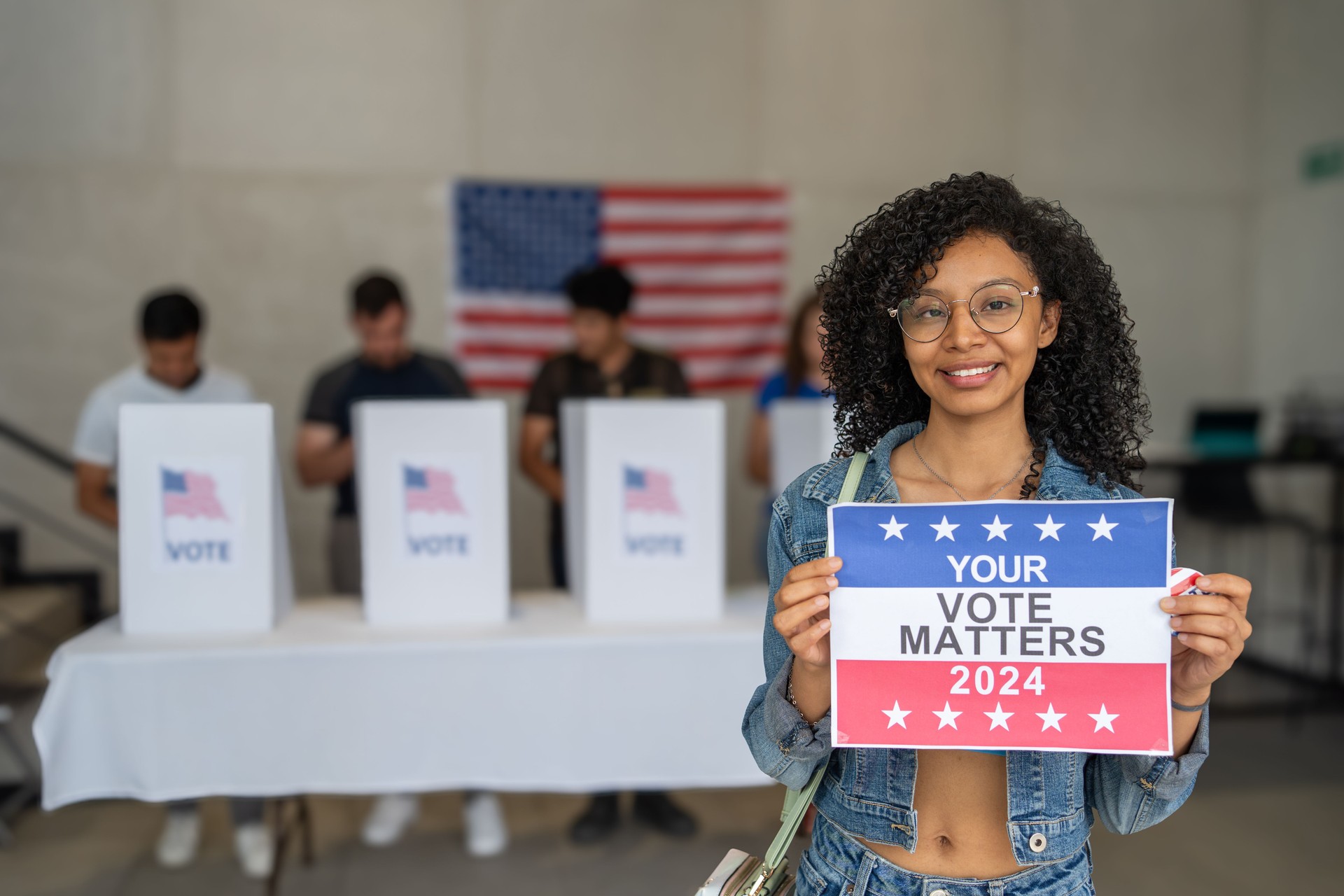 African American woman smiles holding a 'Your Vote Matters 2024' sign at a US election polling station. In the background, a diverse group of people votes in voting booths, democracy in America